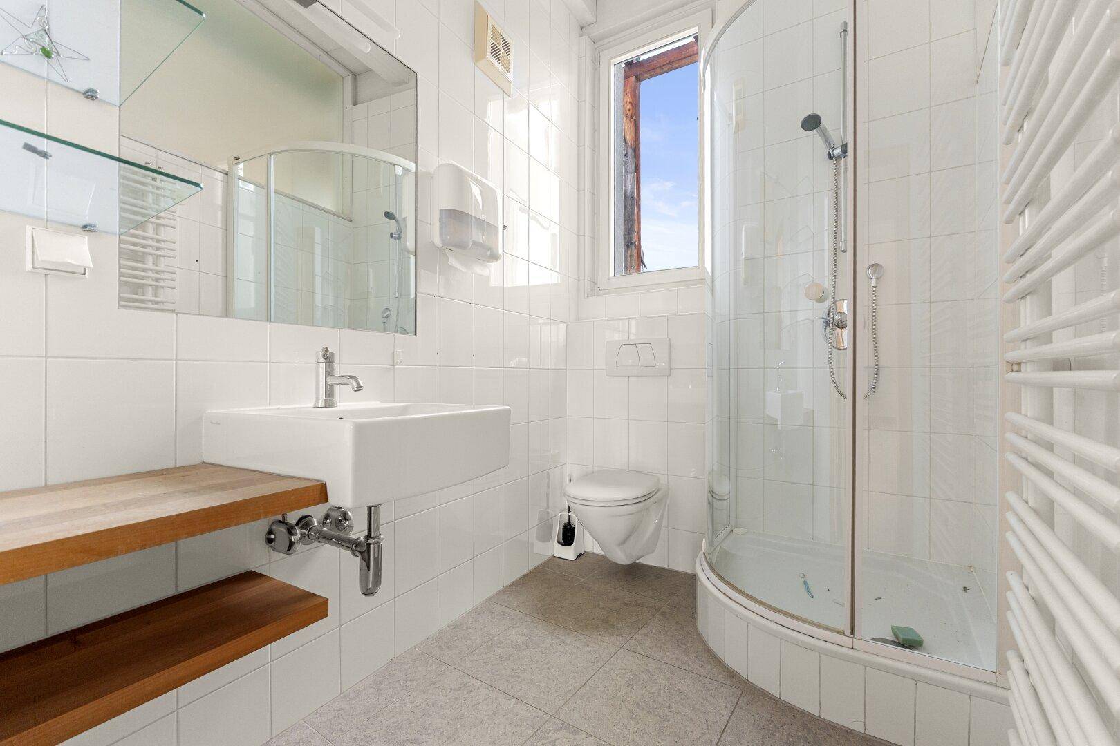 Modernes Badezimmer mit weißen Fliesen, Duschkabine, Waschbecken und Handtuchheizkörper, bietet ein Fenster.