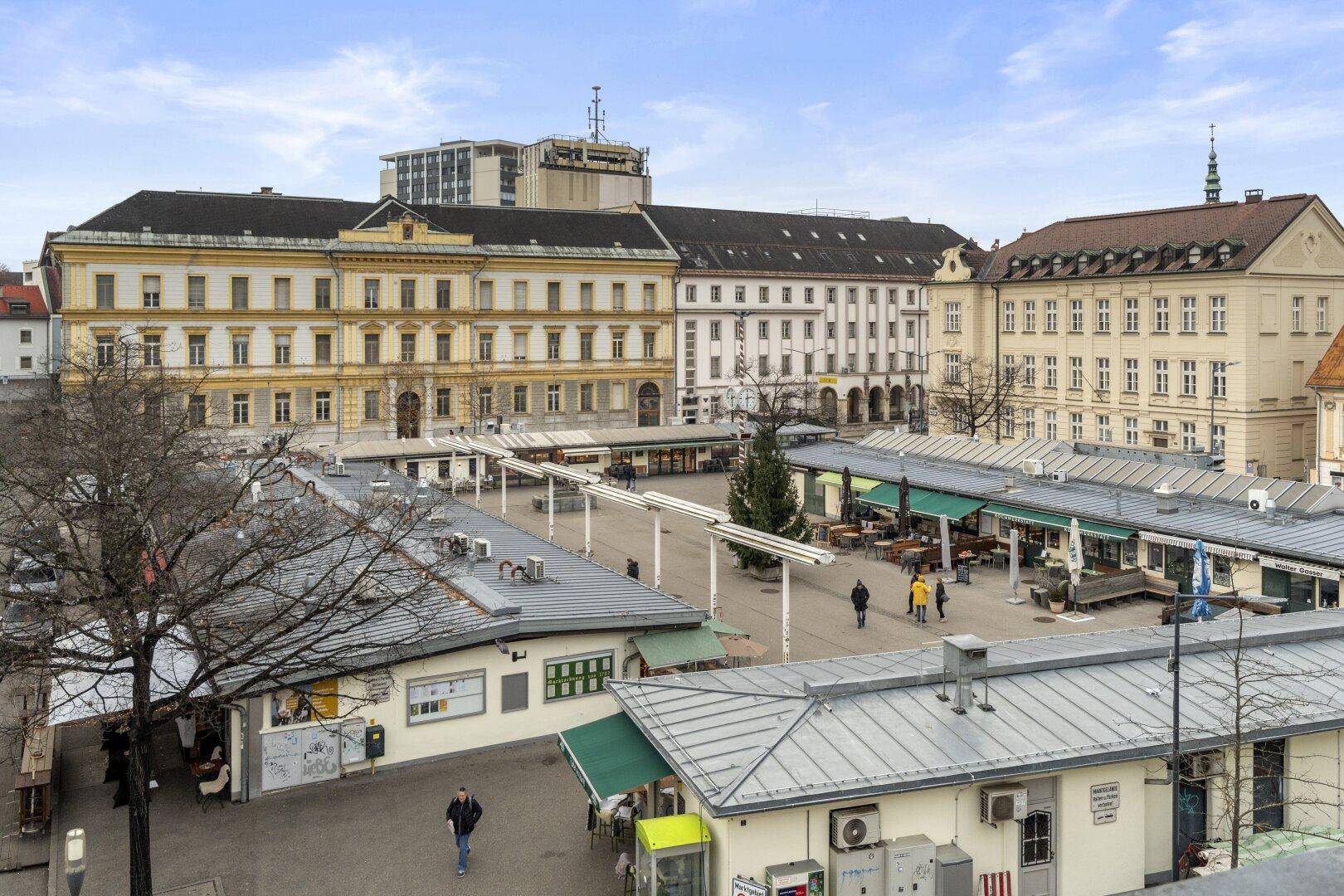 Panoramablick über einen belebten Stadtplatz mit historischen Gebäuden und Marktständen unter blauem Himmel.