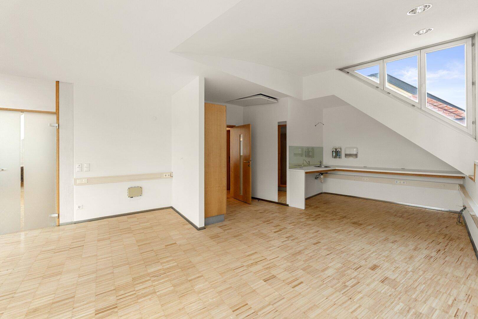 Heller Büroraum mit Holzboden, Dachschrägen und einer kleinen Küchenzeile, ideal für Pausen.