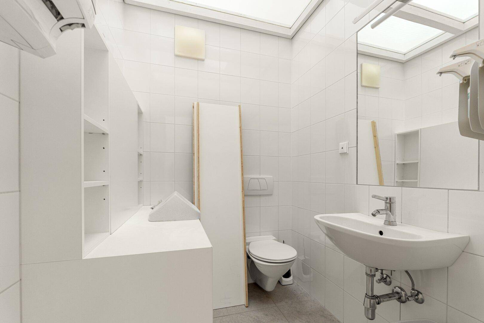 Helles Badezimmer mit weißen Fliesen, Waschbecken und Toilette, bietet Stauraum und Oberlichter.