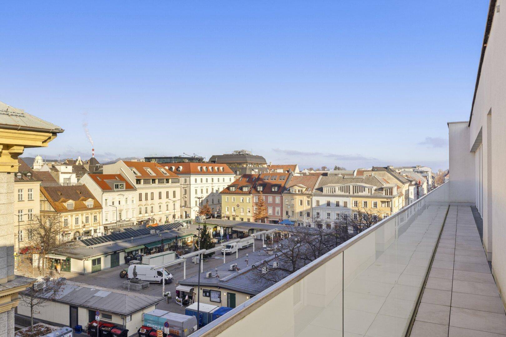 Lange Dachterrasse mit Glasgeländer und Blick auf die belebte Stadt und historische Gebäude.