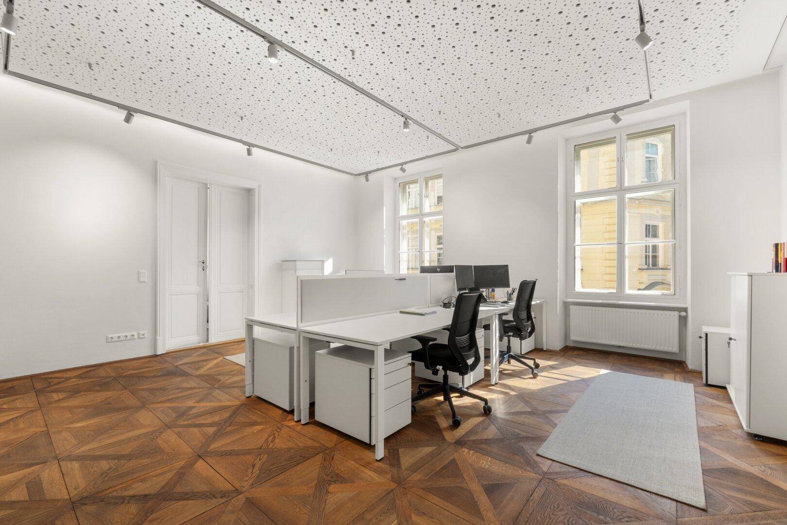 Offener Bürobereich mit mehreren Arbeitsplätzen, großen Fenstern und elegantem Fischgrätparkettboden.