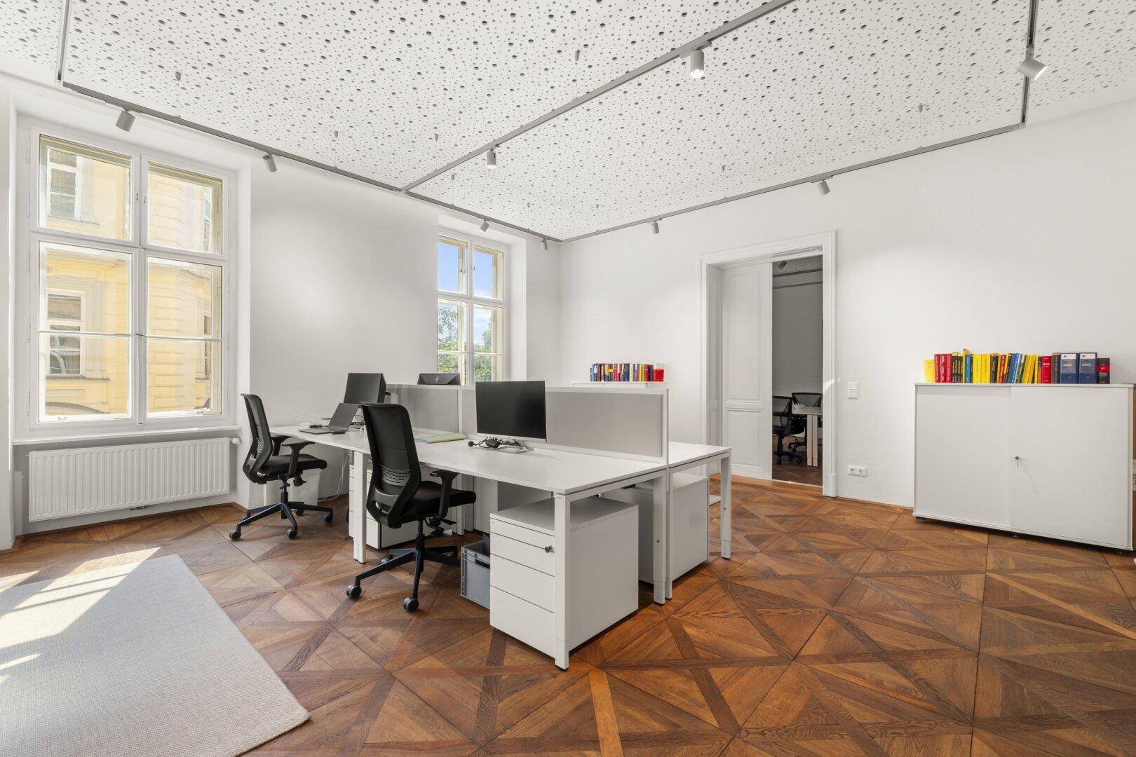 Bürofläche mit mehreren Schreibtischen, Büchern in Regalen und einem hellen Ambiente durch große Fenster.