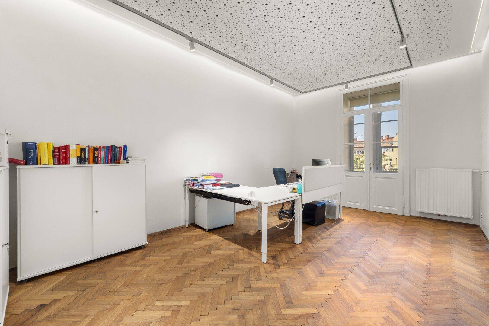 Helles Einzelbüro mit Schreibtisch, Aktenschrank, Fischgrätparkett und hohen Doppeltüren.