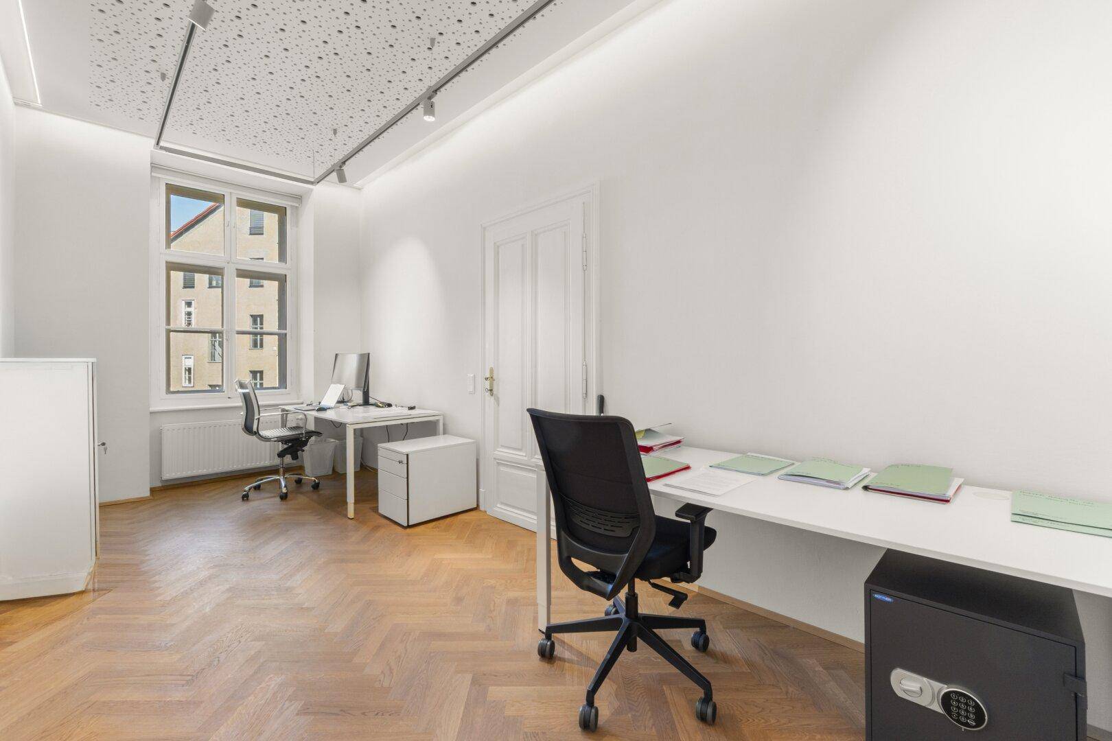 Büroraum mit zwei Arbeitsplätzen, einem Safe, Fischgrätparkett und einem großen Fenster.