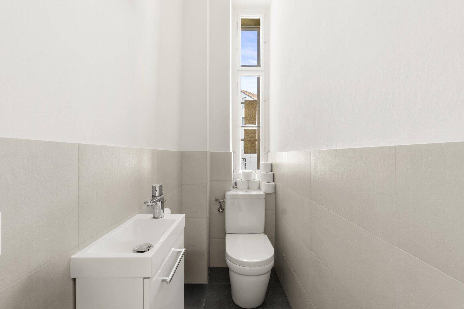 Schmales Gäste-WC mit modernem Waschbecken, Toilette und einem hohen, schmalen Fenster.