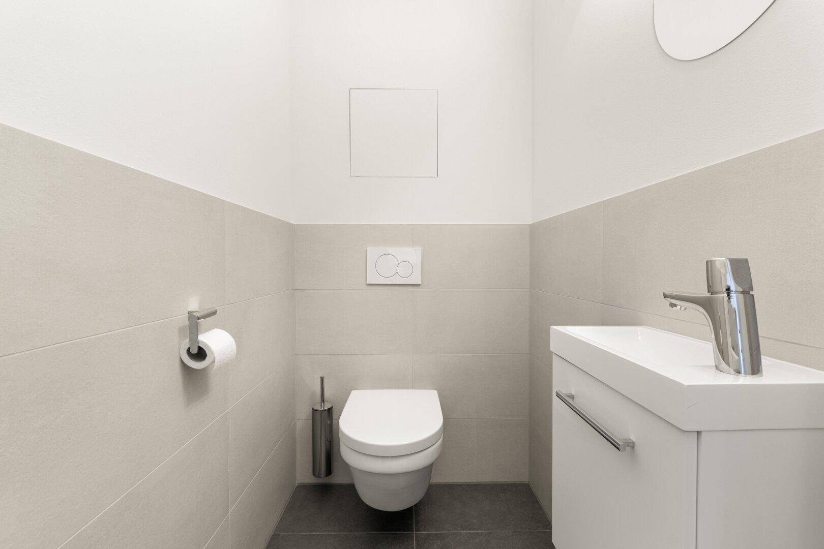 Modernes Gäste-WC mit Wand-WC, kleinem Waschbecken und hellen Fliesen an den Wänden.