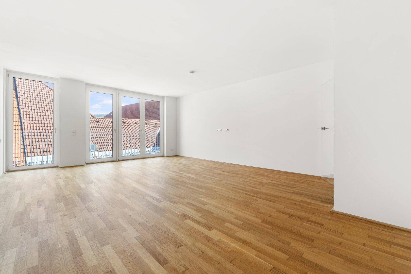 Heller, leerer Raum mit Eichenparkettboden und großen Fenstern mit Blick auf Dächer.
