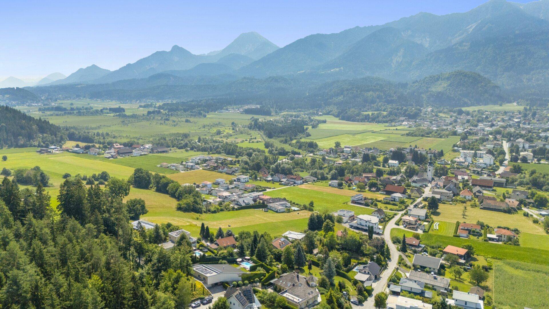 Weitläufige Luftaufnahme einer ländlichen Siedlung mit grünen Feldern und majestätischen Bergen am Horizont.