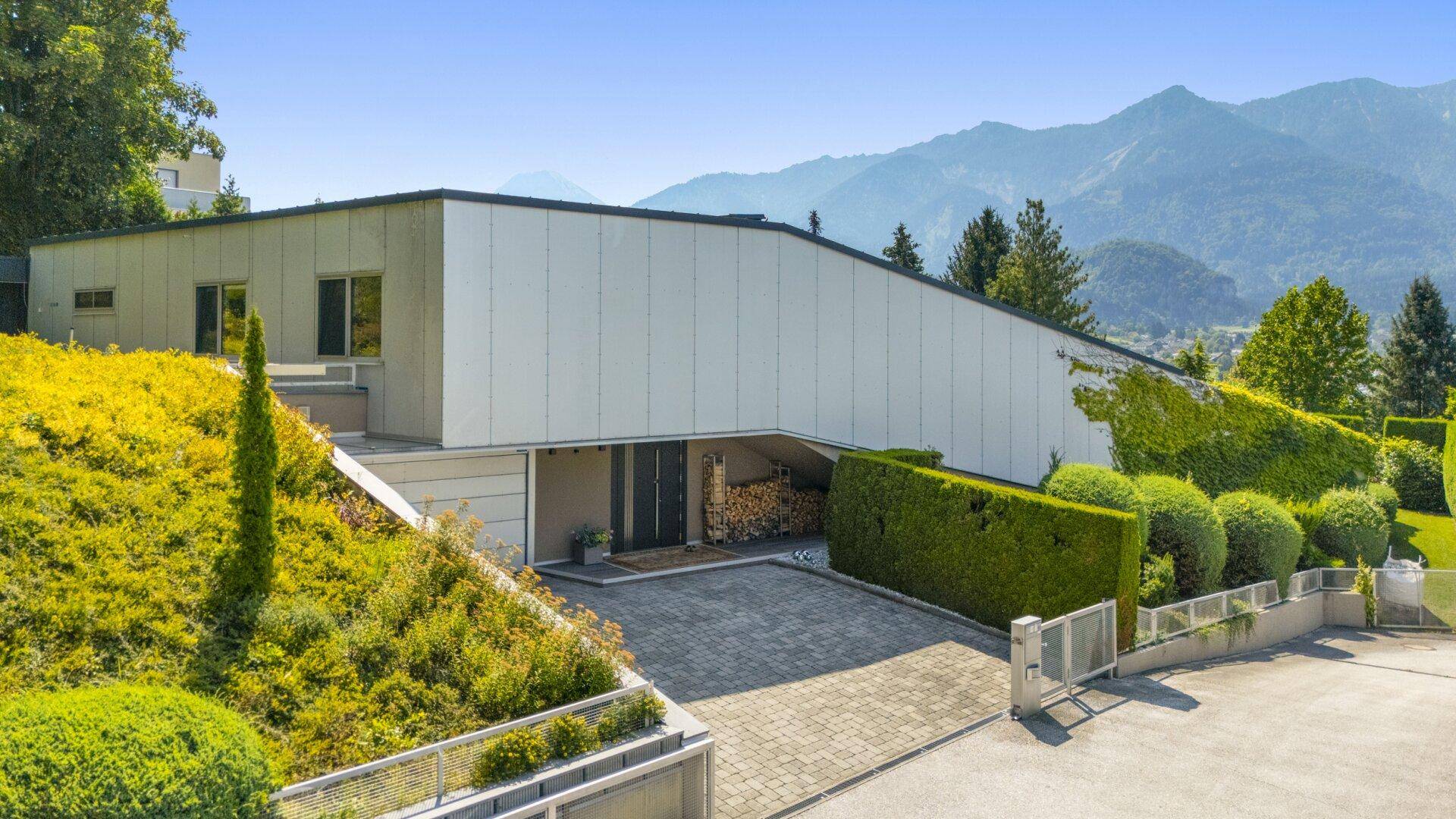 Moderne Architektur mit weißer Fassade, umgeben von grüner Landschaft und Bergen im Hintergrund.