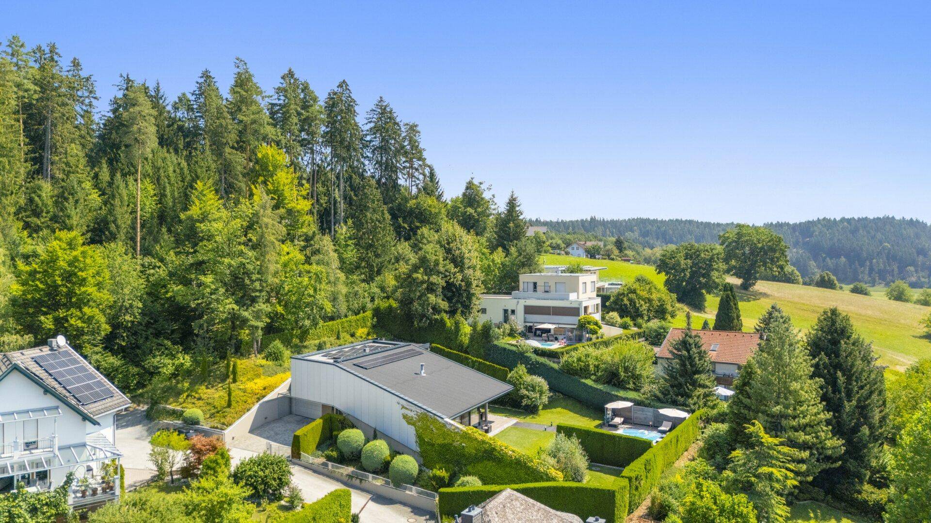 Luftaufnahme eines modernen Hauses mit Pool, umgeben von üppigem Grün und Wald am Hang.