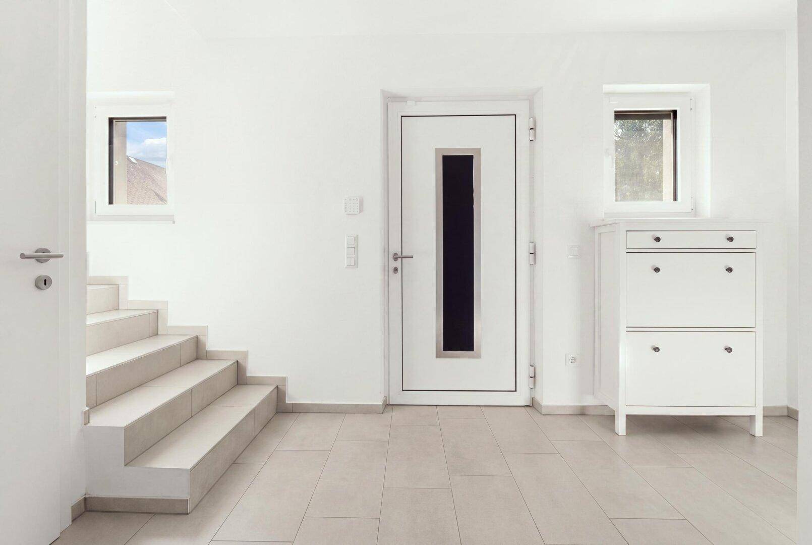 Heller Eingangsbereich mit Fliesenboden, Treppe und moderner Haustür mit Glaseinsatz.
