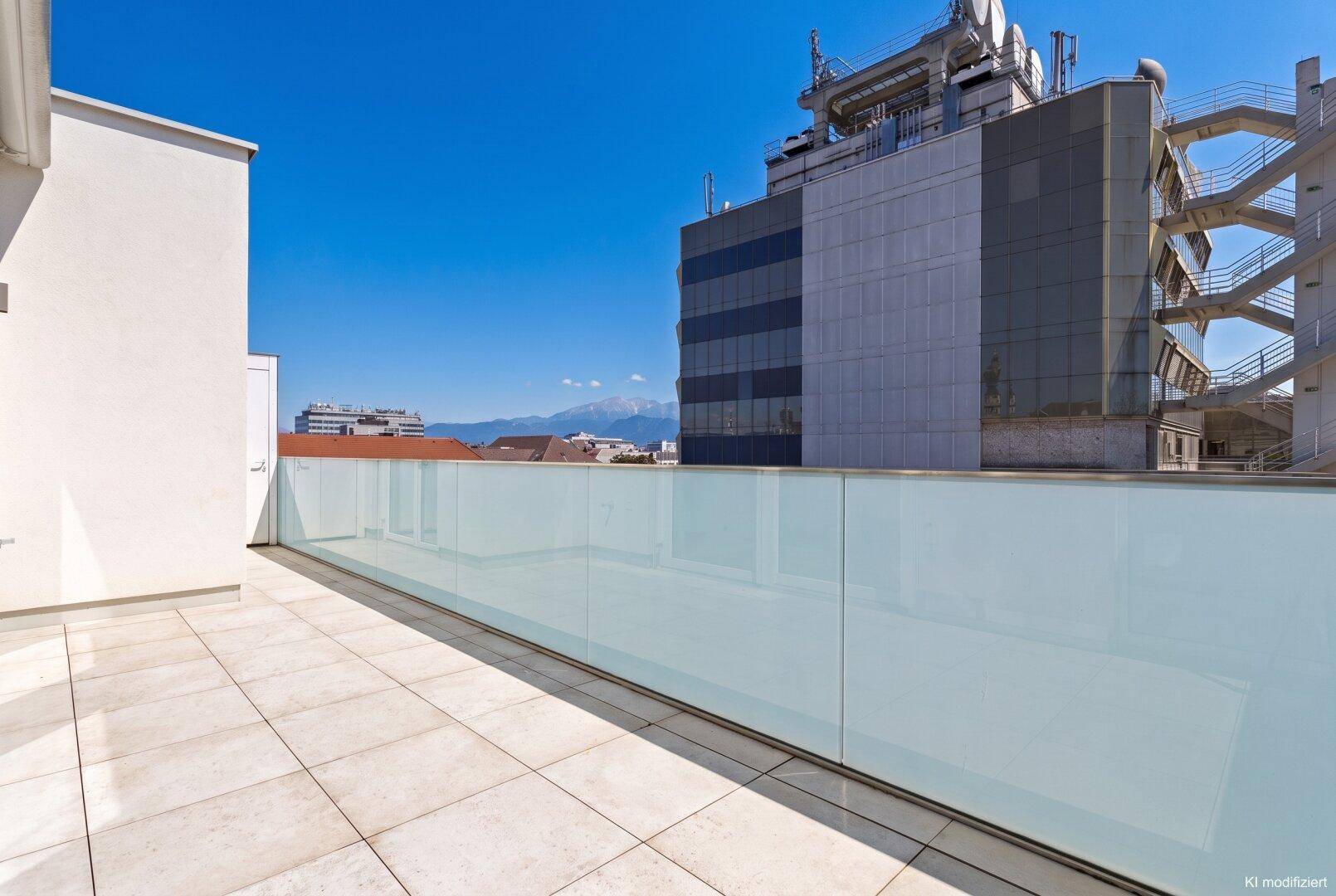 Geräumige Dachterrasse mit hellen Fliesen, Glasgeländer und Blick auf die Stadt und Berge in der Ferne.