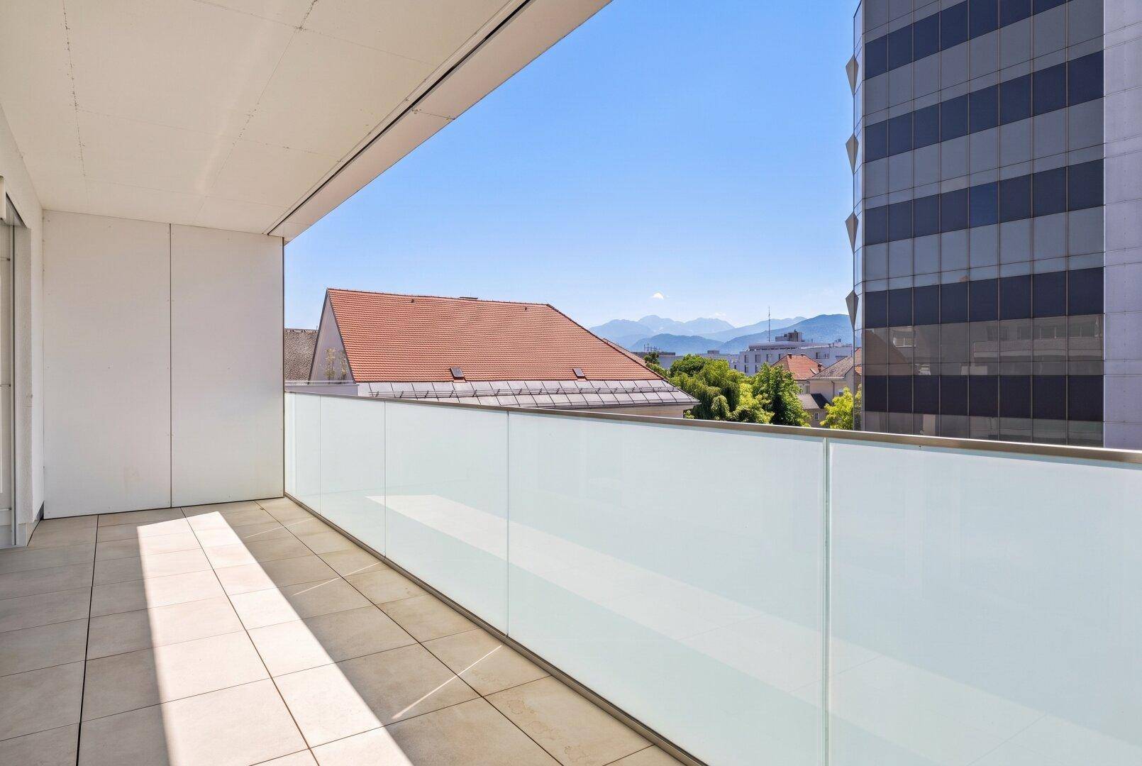 Sonniger Balkon mit zeitgemäßer Glasbrüstung, gefliestem Boden und Panoramablick auf die Stadt und die Alpen.