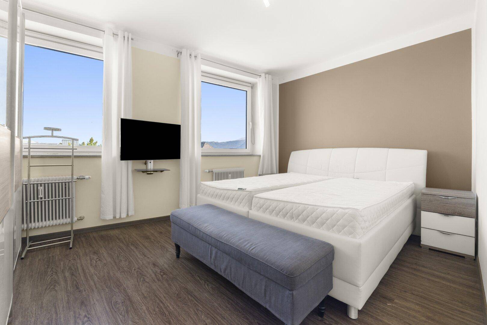 Helles Schlafzimmer mit Doppelbett, zwei Fenstern und dunklem Holzboden.