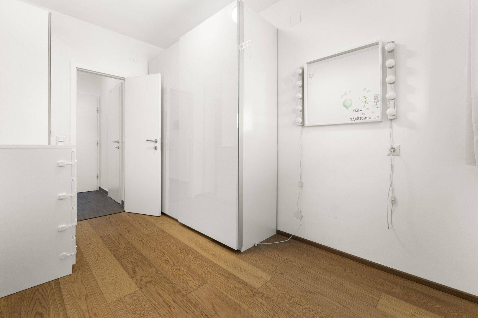 Schlafzimmer mit weißen Kleiderschränken, Holzboden und beleuchtetem Schminkspiegel.