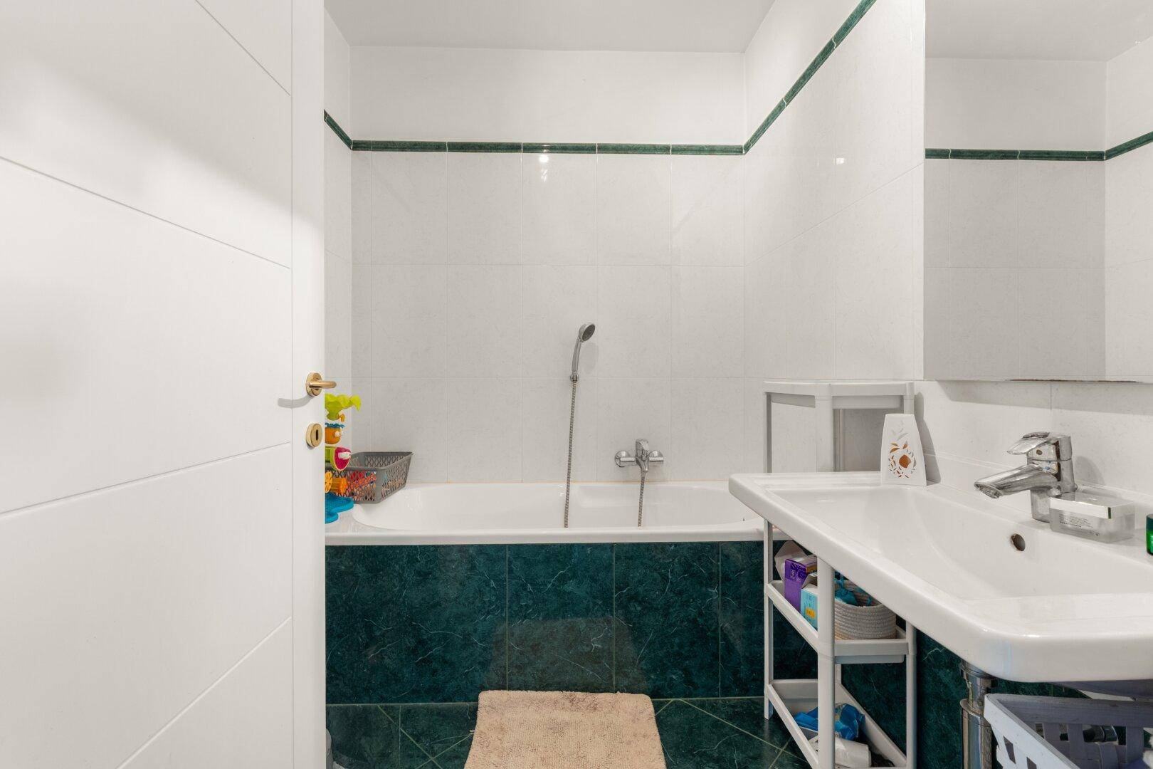 Badezimmer mit Badewanne, Waschbecken und weißen Fliesen mit grünen Akzenten.