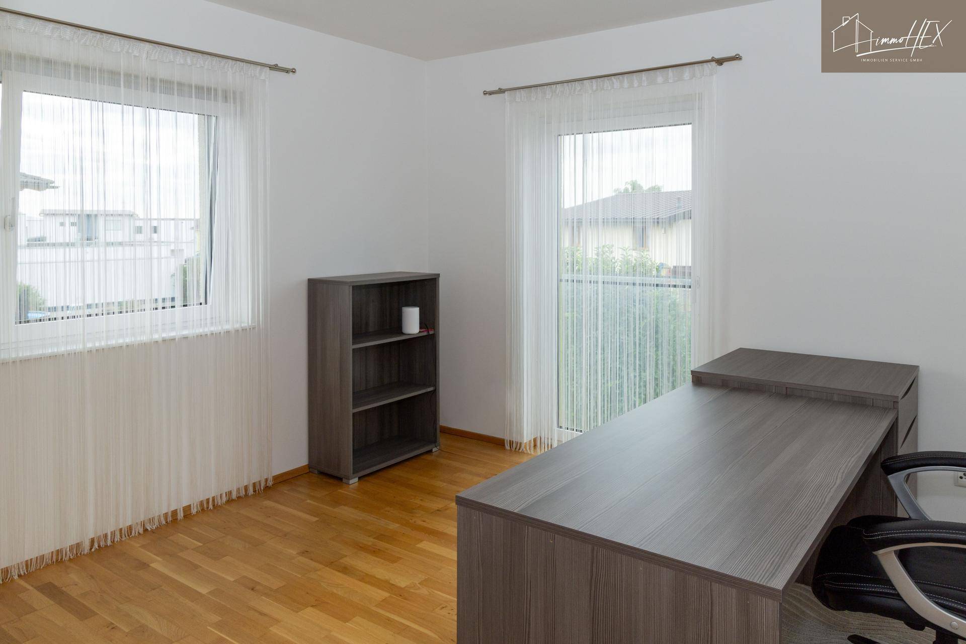 Helles Arbeitszimmer mit Holzboden, großem Fenster und Balkonzugang, ausgestattet mit Schreibtisch und Regal.