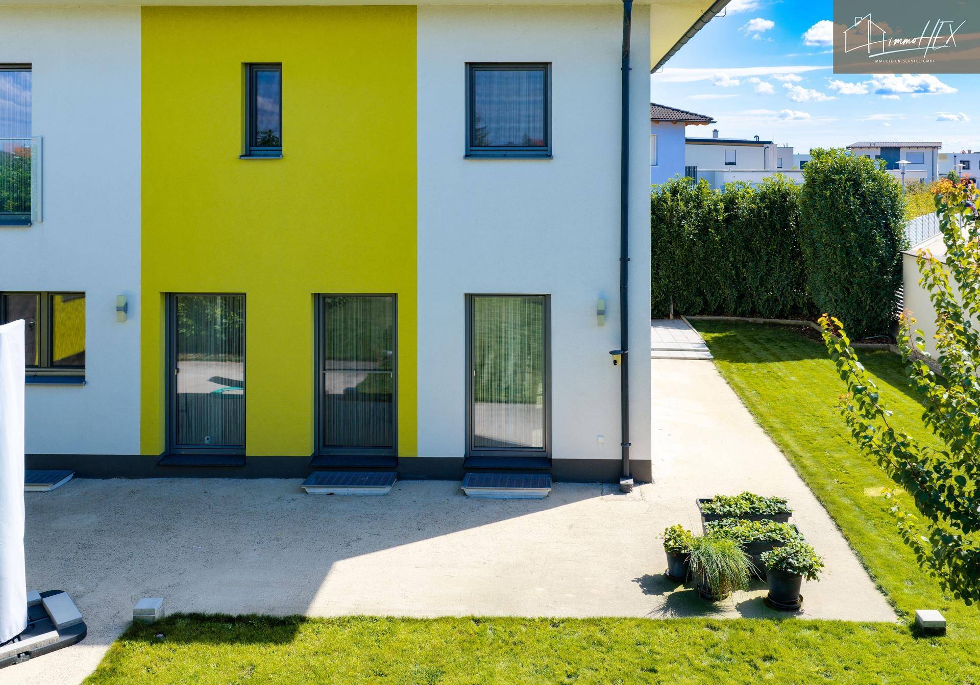 Rückseite des Hauses mit Terrassenzugang, Rasenfläche und hohen Hecken für Privatsphäre.