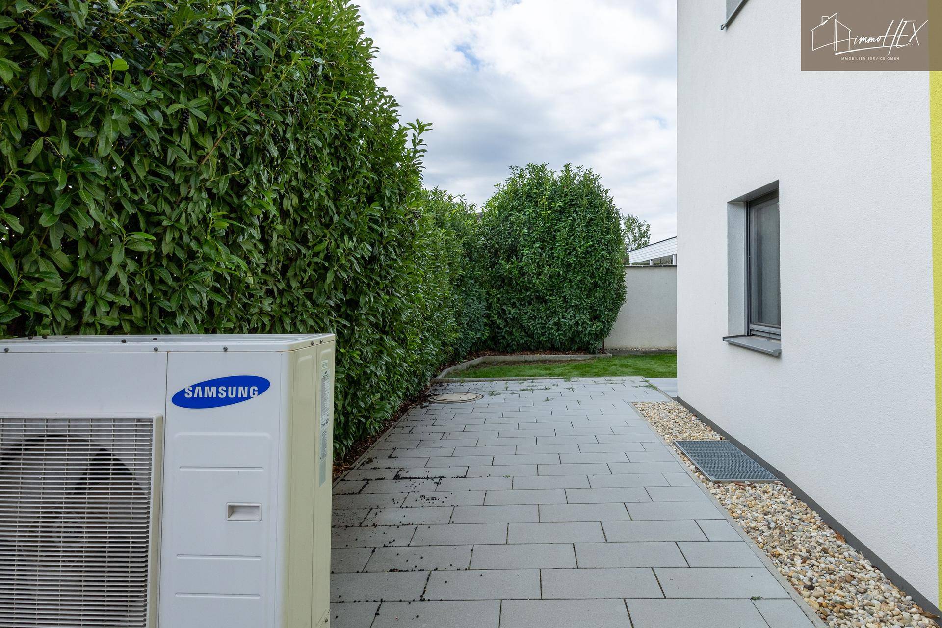 Außenansicht des Hauses mit Wärmepumpe und gepflastertem Bereich neben einer Hecke.