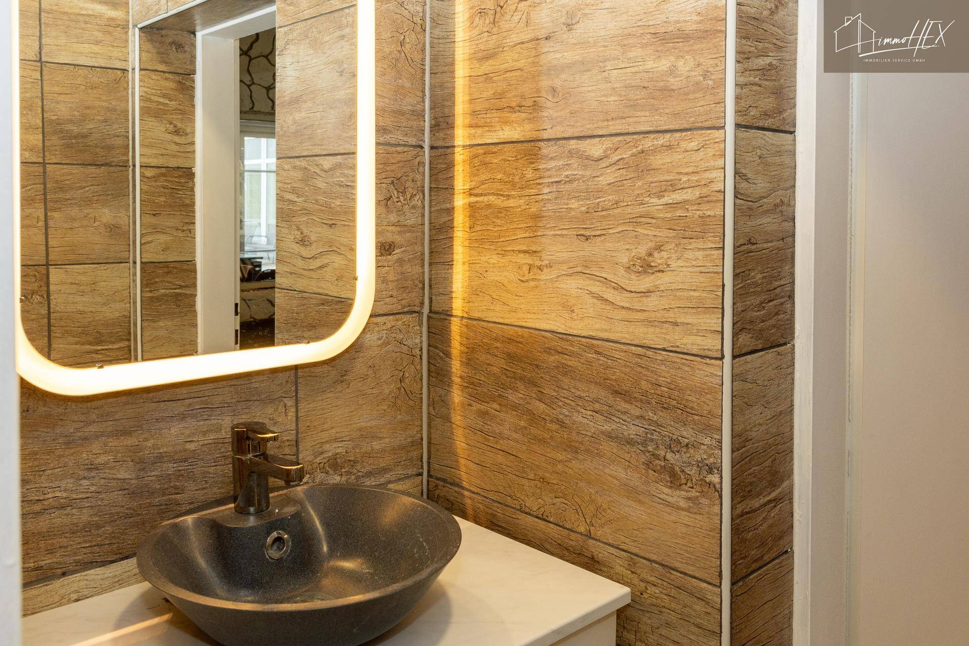 Modernes Waschbecken mit beleuchtetem Spiegel und holzgemusterten Fliesen.