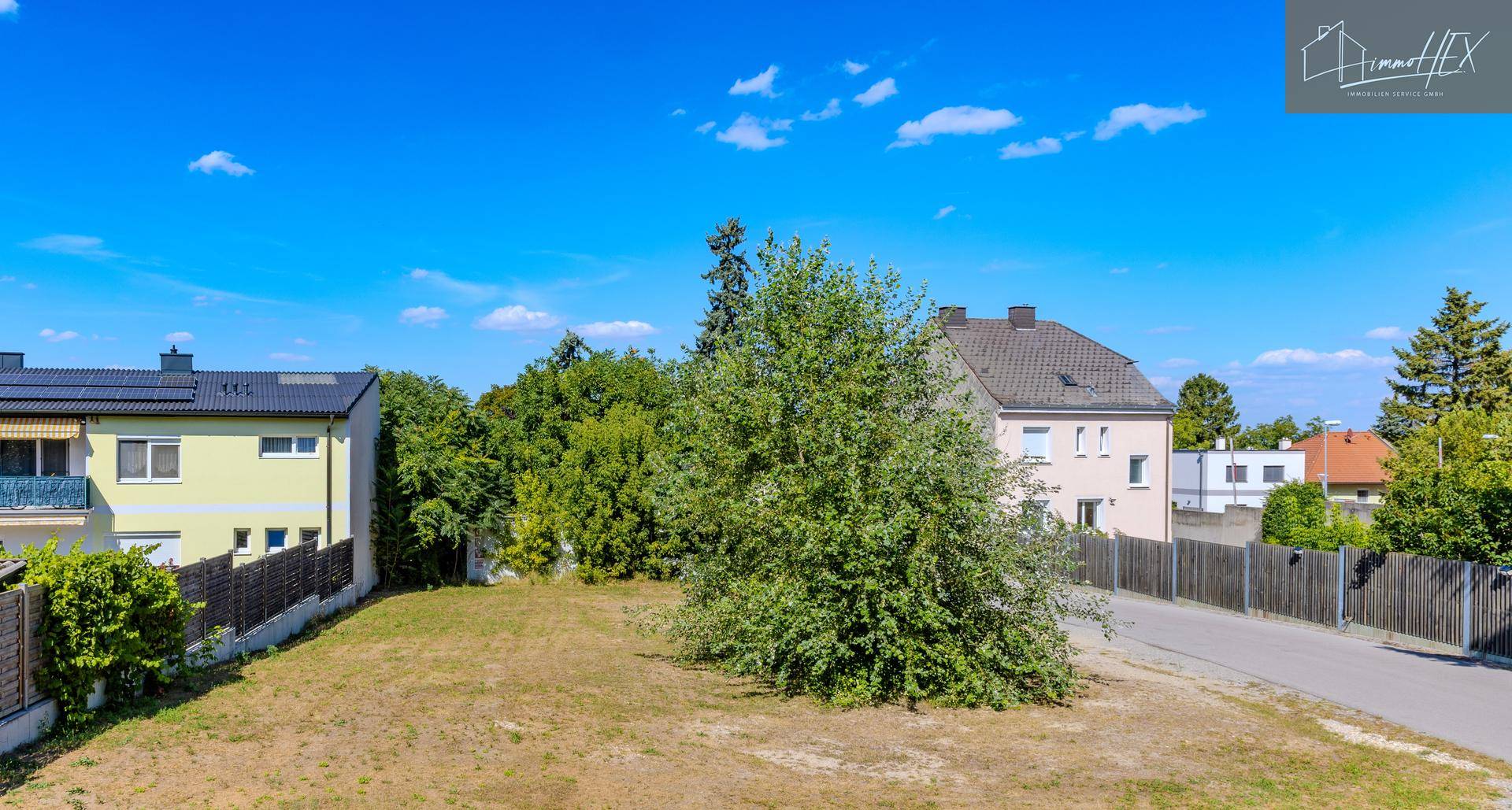 Blick auf ein unbebautes Grundstück mit einem Baum, umgeben von Wohnhäusern und einer ruhigen Straße.