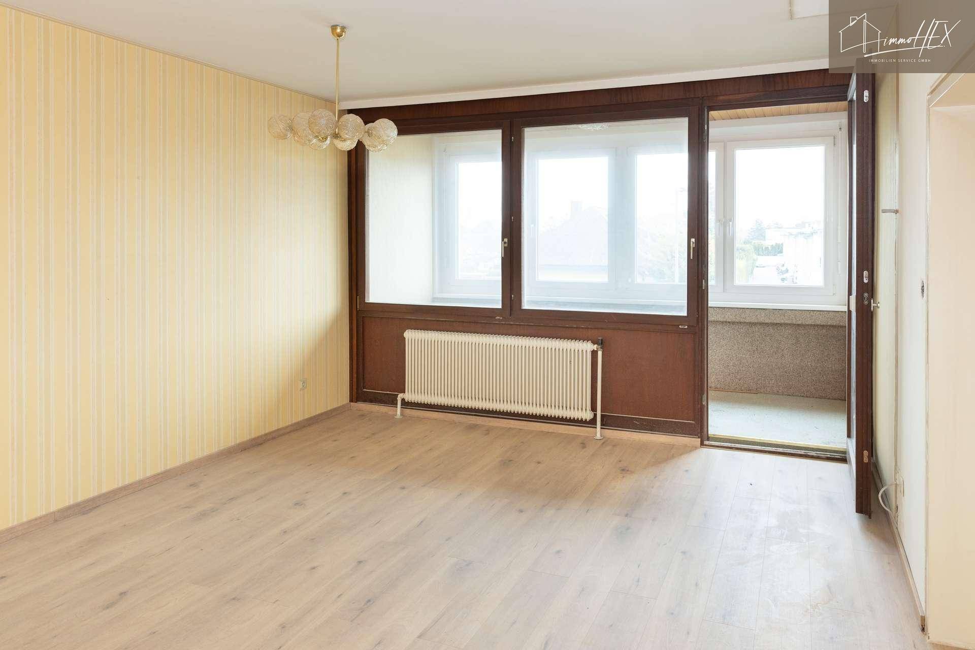 Geräumiges Wohnzimmer mit älterer Tapete, dunklen Fensterrahmen und Zugang zu einem kleinen Balkon.