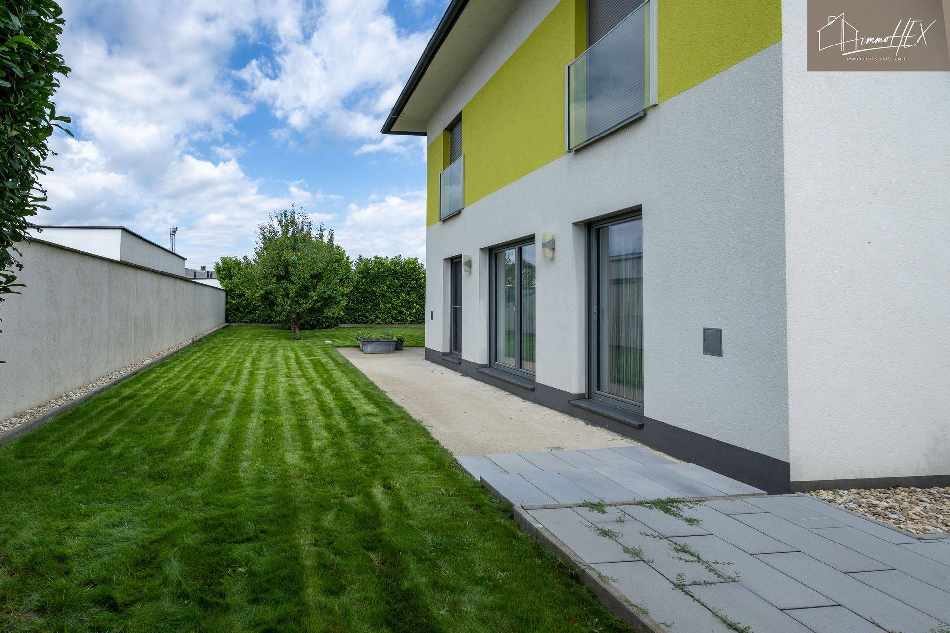 Langer, schmaler Gartenbereich mit Rasen und einer gepflasterten Terrasse entlang der Hauswand.