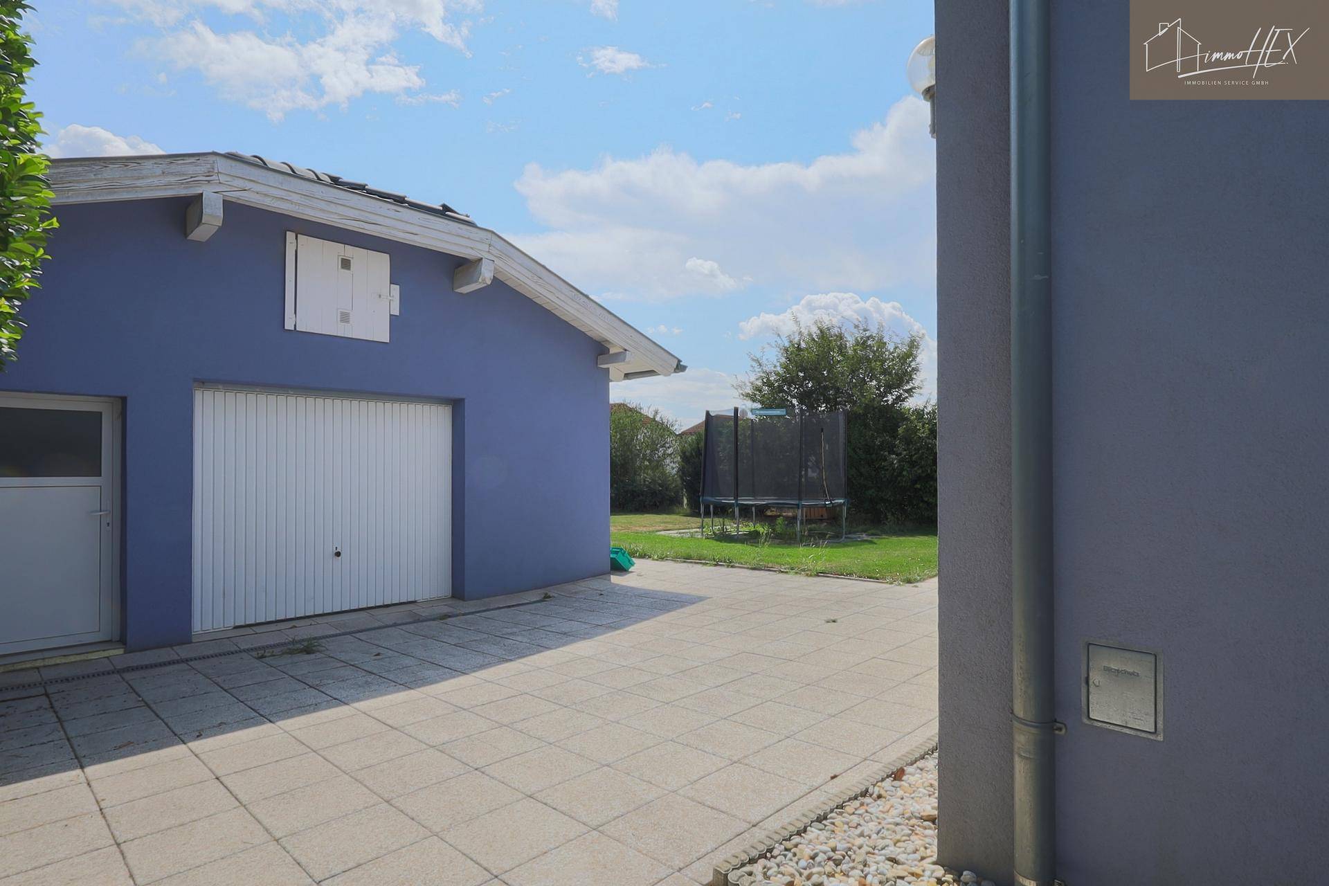 Seitenansicht des Garagengebäudes mit Einzeltor, gepflastertem Bereich und Trampolin im Hintergrund.