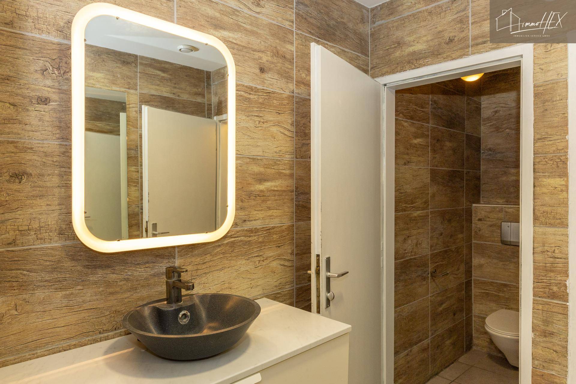Heller Toilettenbereich mit modernem Waschbecken, beleuchtetem Spiegel und holzgemusterten Fliesen.