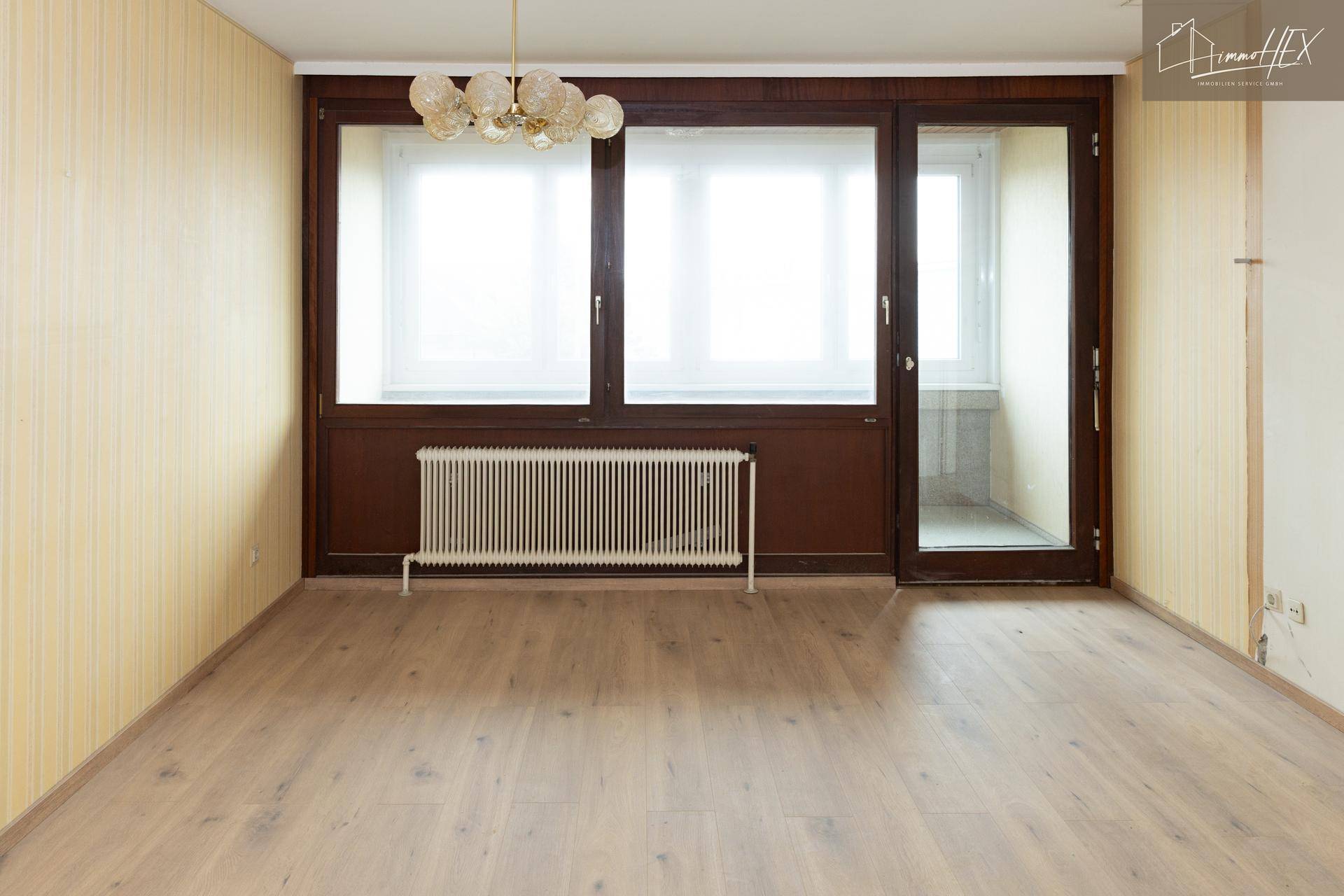 Leeres Wohnzimmer mit gestreifter Tapete, dunklen Holzfenstern und Zugang zu einem kleinen Balkon.