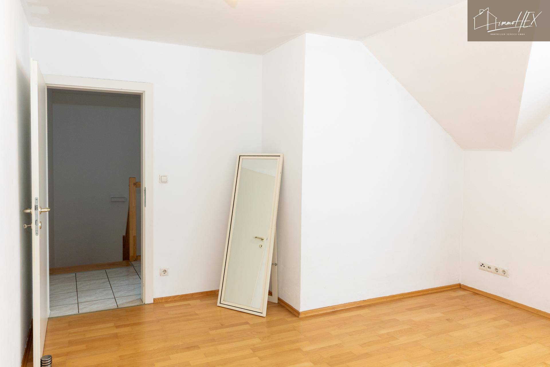 Helles Zimmer mit Holzboden, weiß gestrichenen Wänden und Zugang zu einem weiteren Bereich.