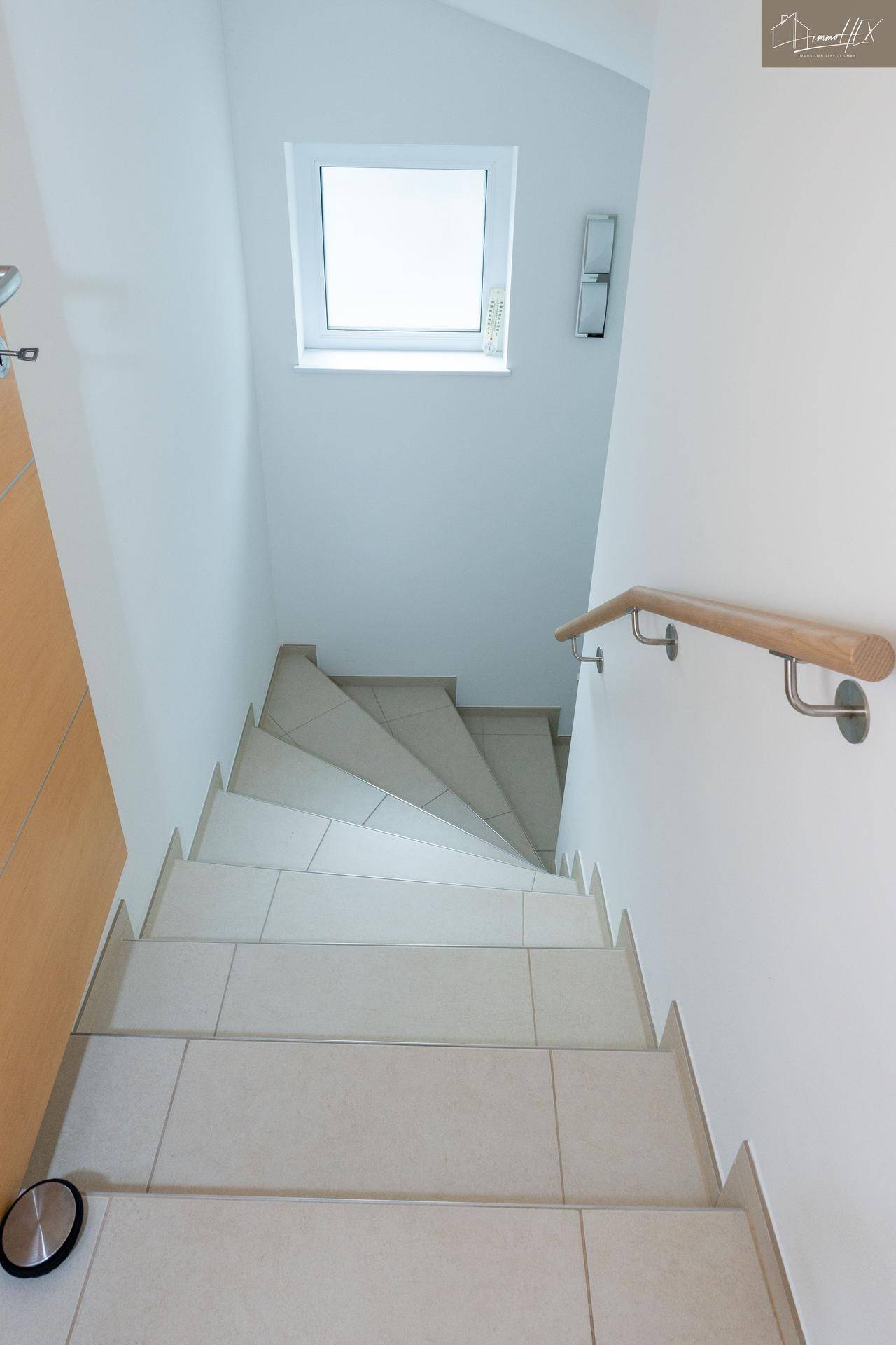 Treppenabgang mit hellen Fliesen und Holzhandlauf, der in den unteren Bereich des Hauses führt.