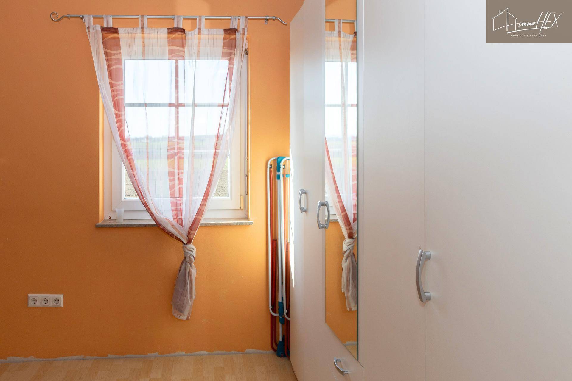 Fenster in einem Zimmer mit orangefarbenen Wänden und einem weißen Kleiderschrank mit Spiegeltür.