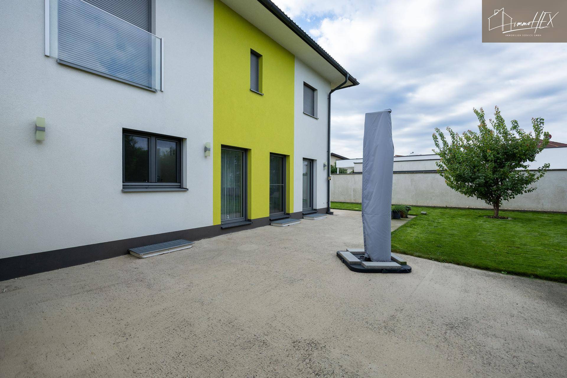 Große gepflasterte Terrasse neben dem modernen Haus mit Rasenfläche und Sonnenschirmständer.