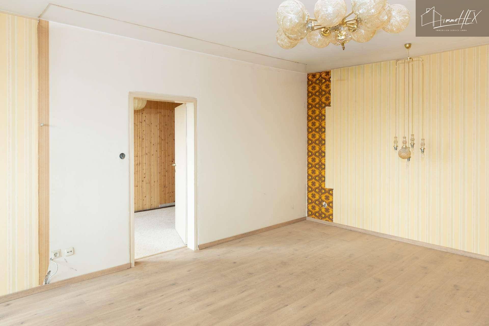 Geräumiges Wohnzimmer mit gemusterter Tapete und offenem Durchgang zu einem weiteren Raum.