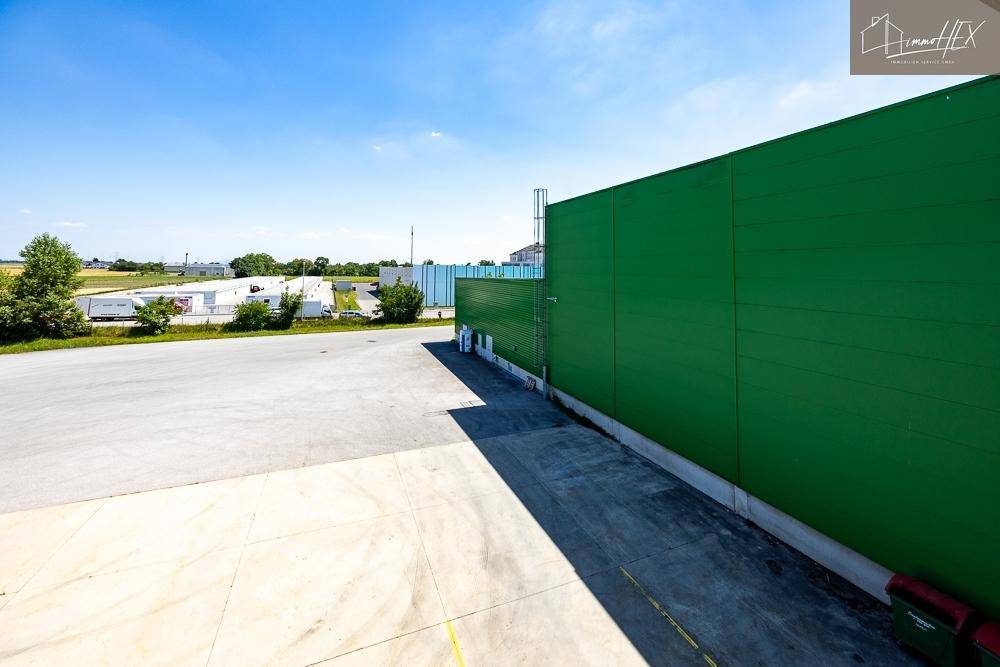 Außenansicht eines grünen Industriegebäudes mit großem Parkplatz unter blauem Himmel.