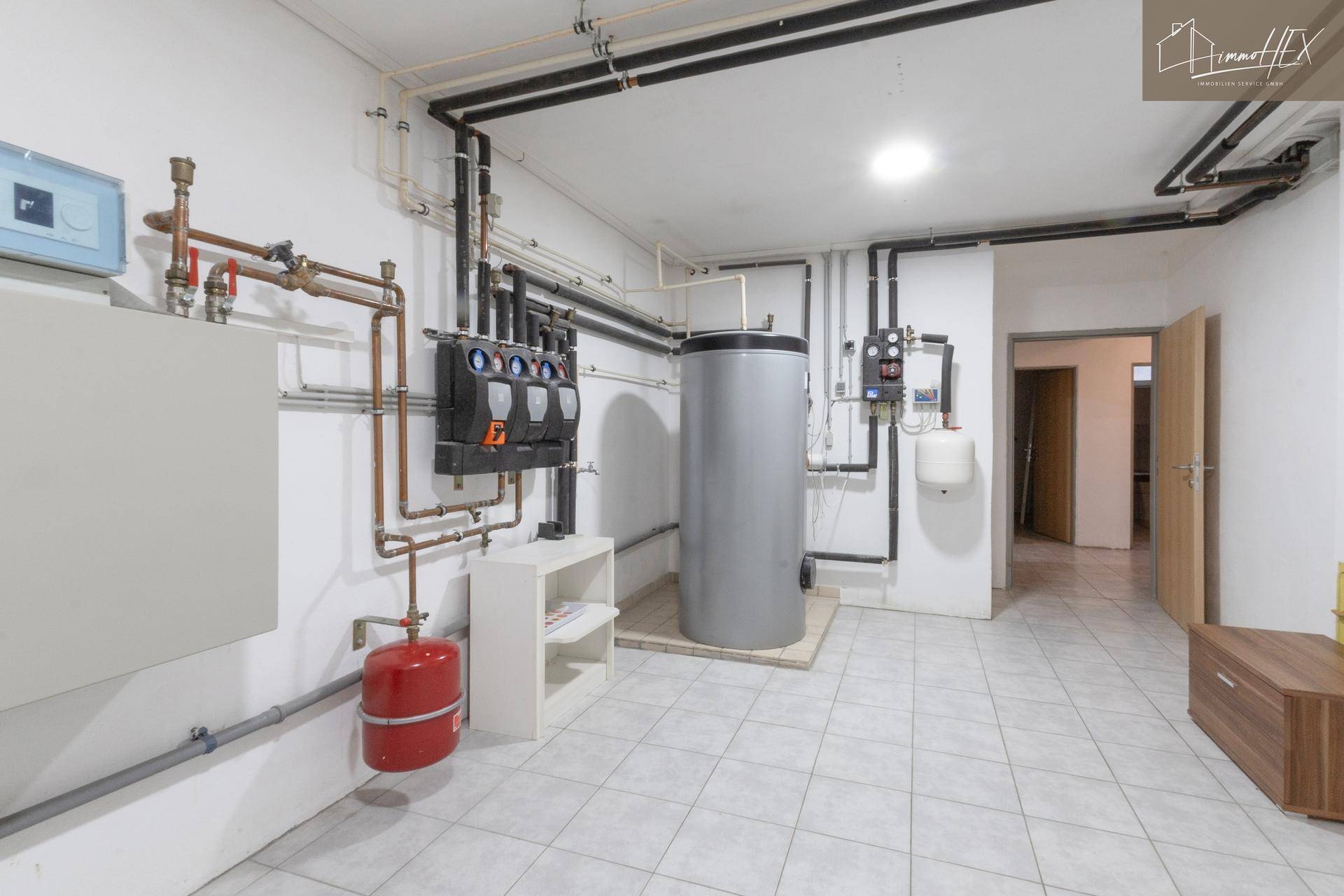 Technikraum im Keller mit Heizungsanlage, Warmwasserspeicher und sichtbaren Leitungen.