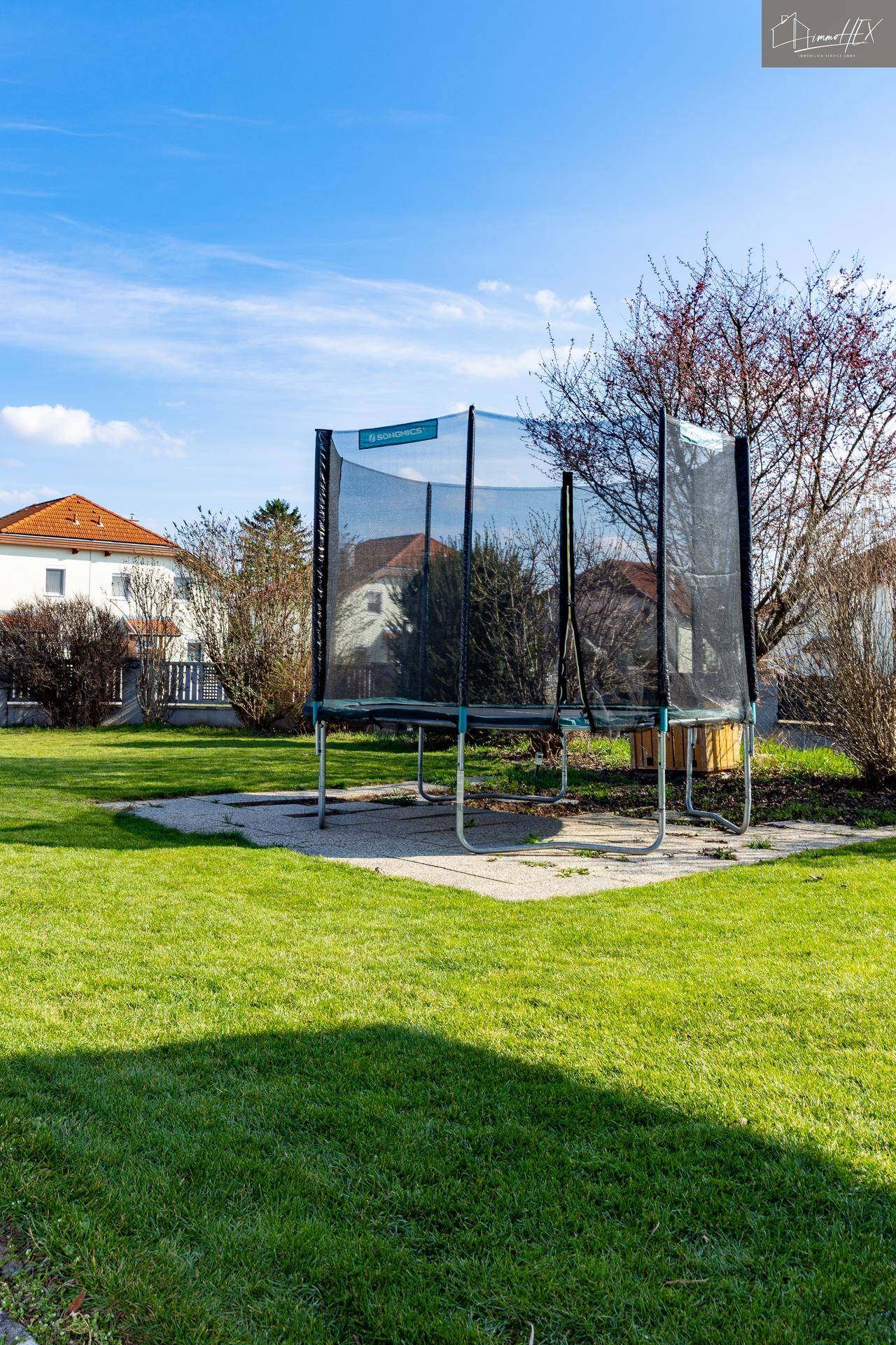 Sonniger Garten mit großem Trampolin und Rasenfläche, umgeben von Bäumen.