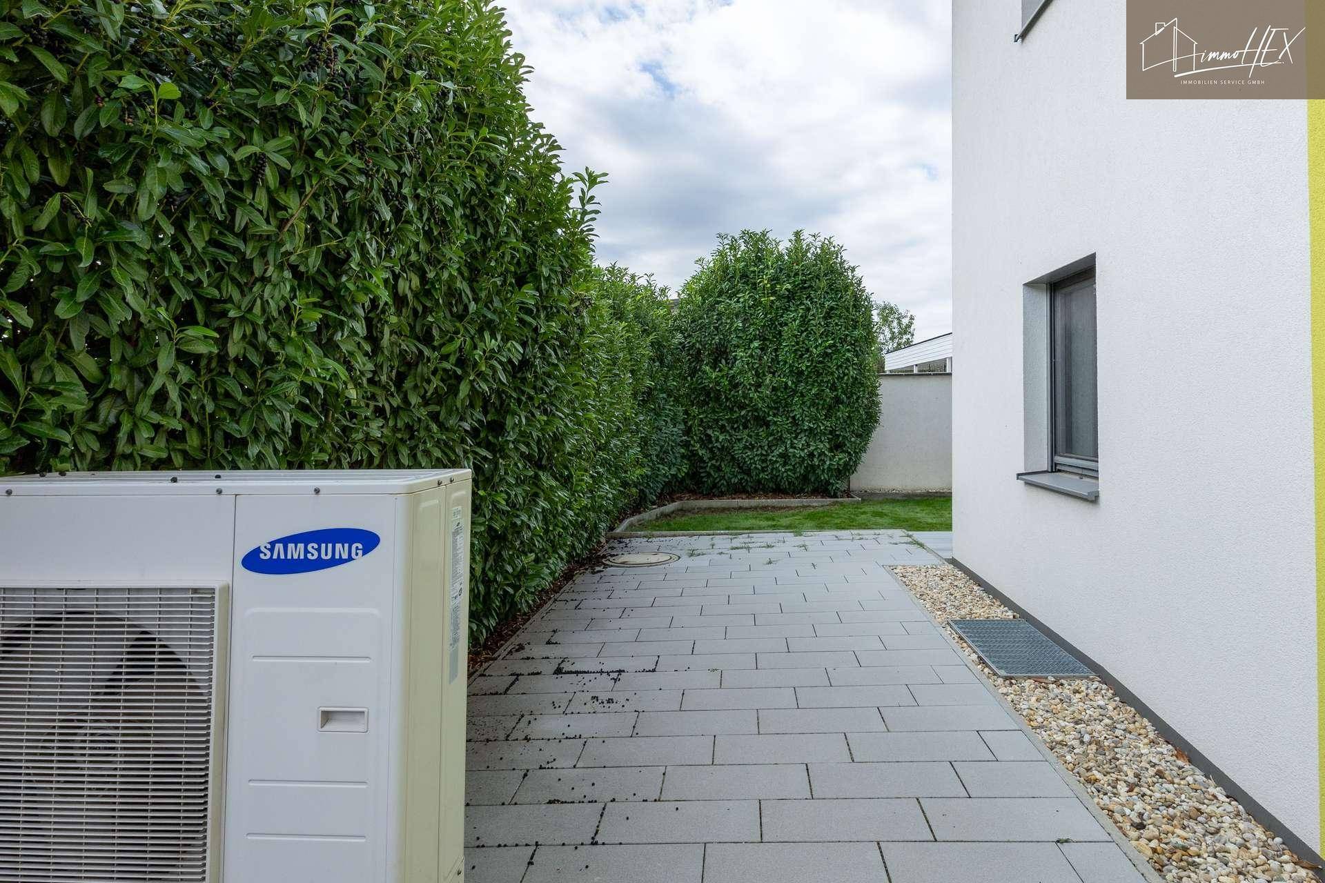 Außenansicht des Hauses mit einer Wärmepumpe und gepflastertem Weg neben der Hecke.