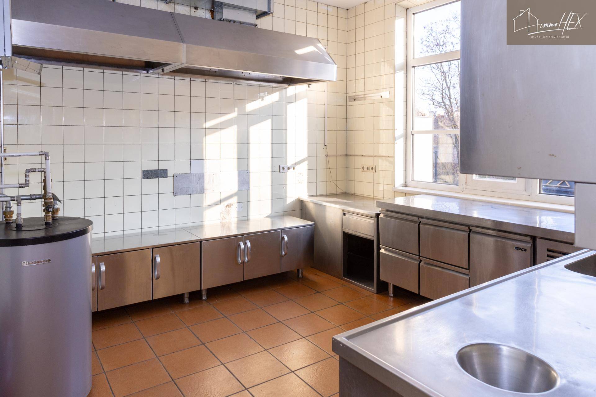 Professionelle Großküche mit Edelstahlgeräten, Fliesenwänden und großem Fenster.