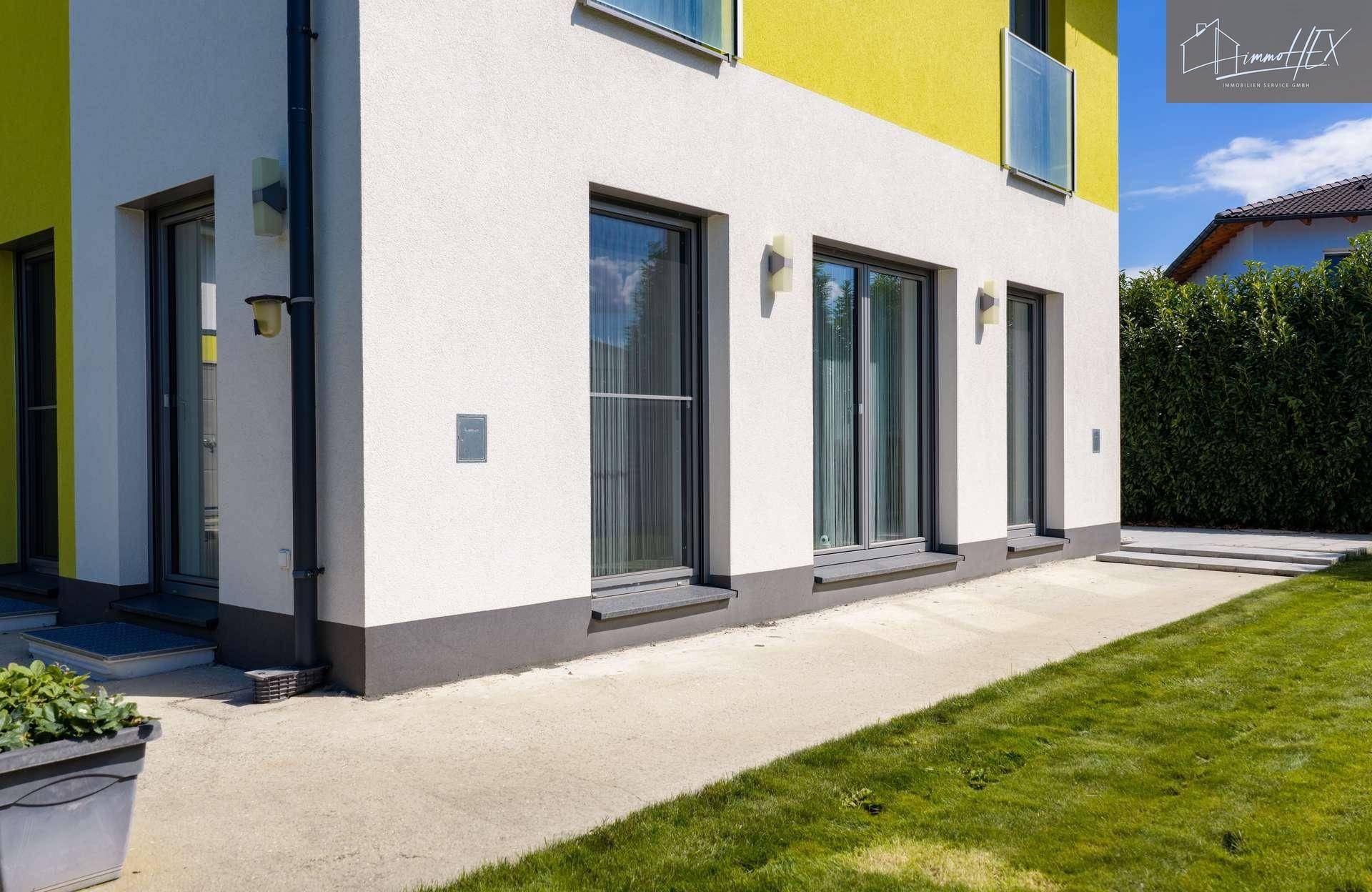 Seitenansicht des Hauses mit großen Fenstern, Zugang zum Garten und gepflegter Rasenfläche.