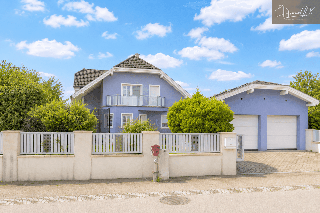 Ansicht des hellblauen Einfamilienhauses und der Doppelgarage von der Straße mit Zaun und Bepflanzung.