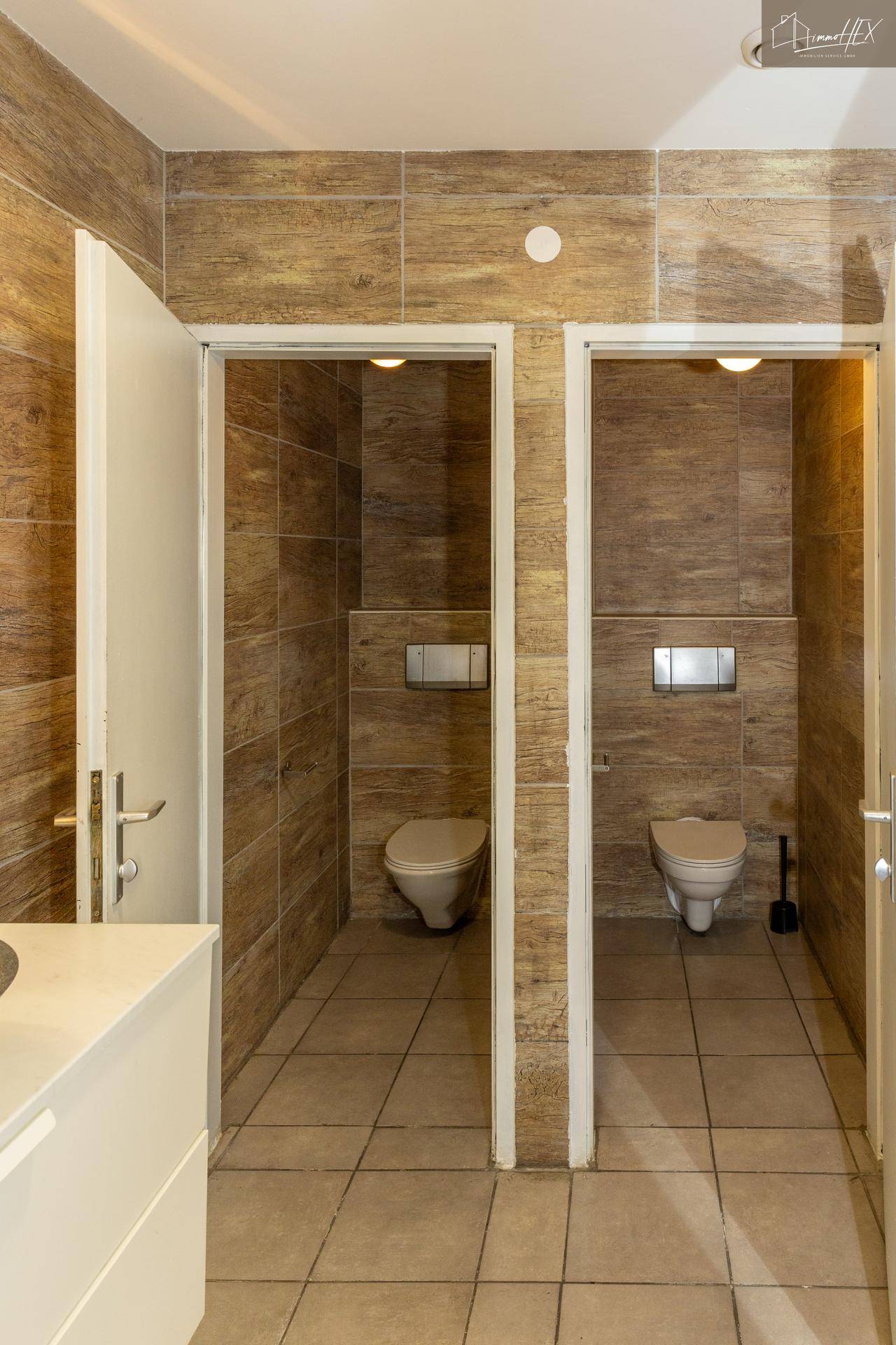Zwei separate Toilettenkabinen mit modernen Wand-WCs und holzgemusterten Fliesen.
