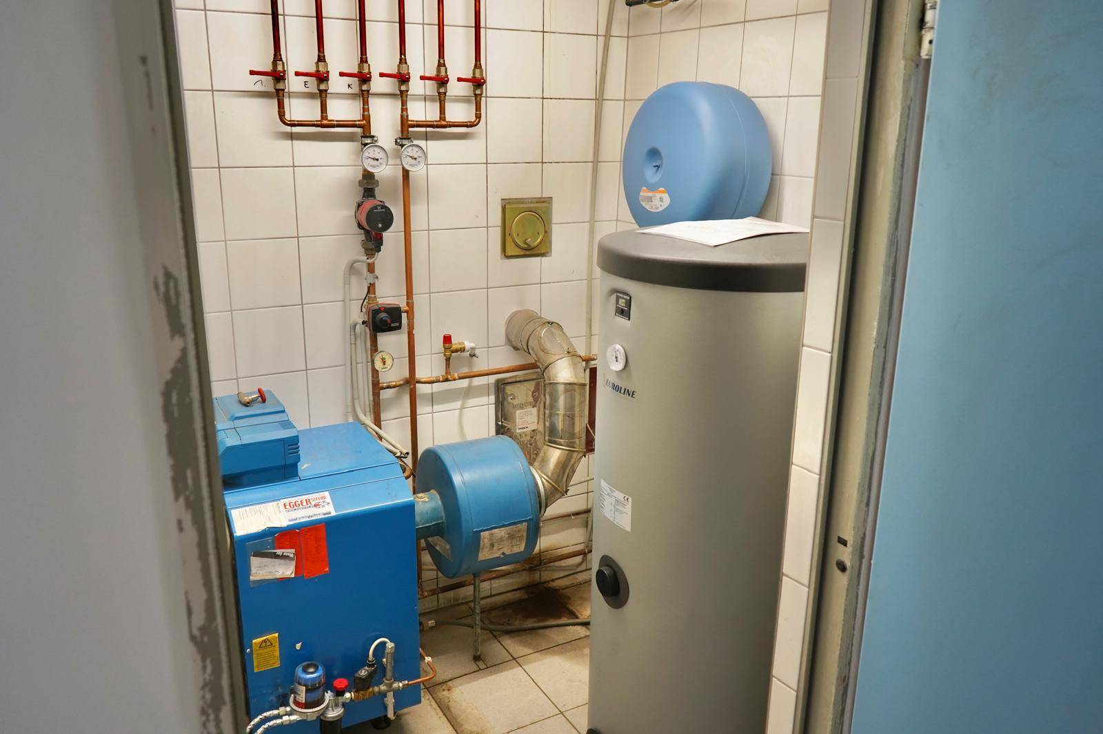 Heizungsraum mit Boiler und Rohrleitungen, gefliesten Wänden und Boden.