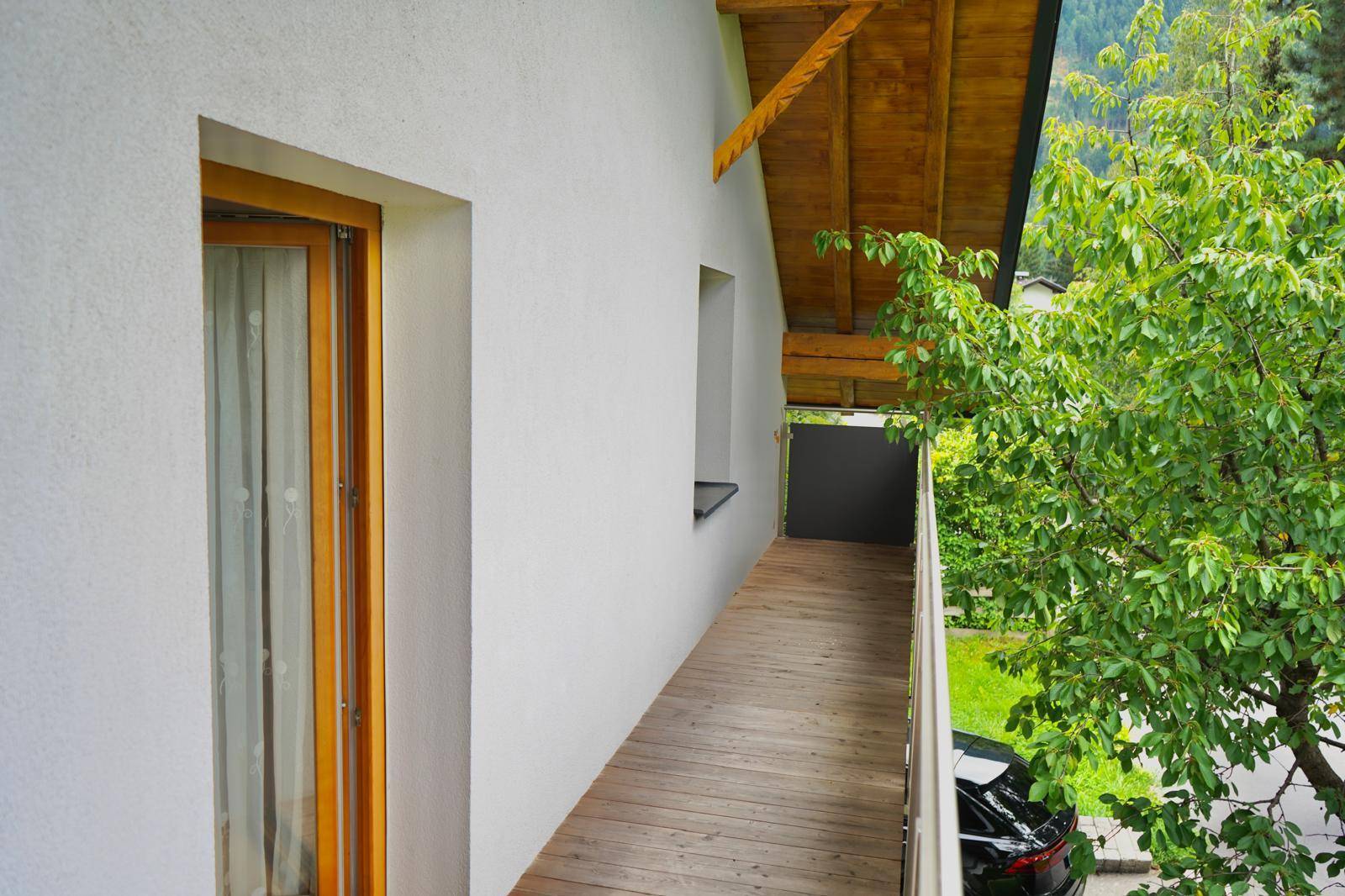 Langer Holzbalkon mit Geländer und Zugang zu den Zimmern, umgeben von Bäumen.