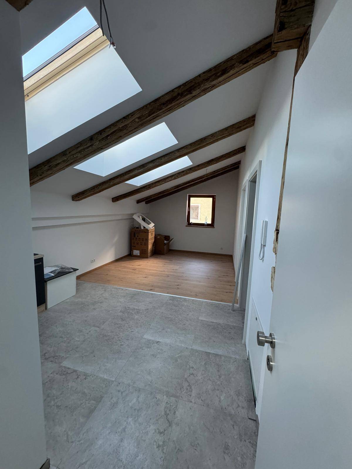 Heller Dachbodenraum mit sichtbaren Holzbalken, Dachfenstern und gemischtem Bodenbelag.