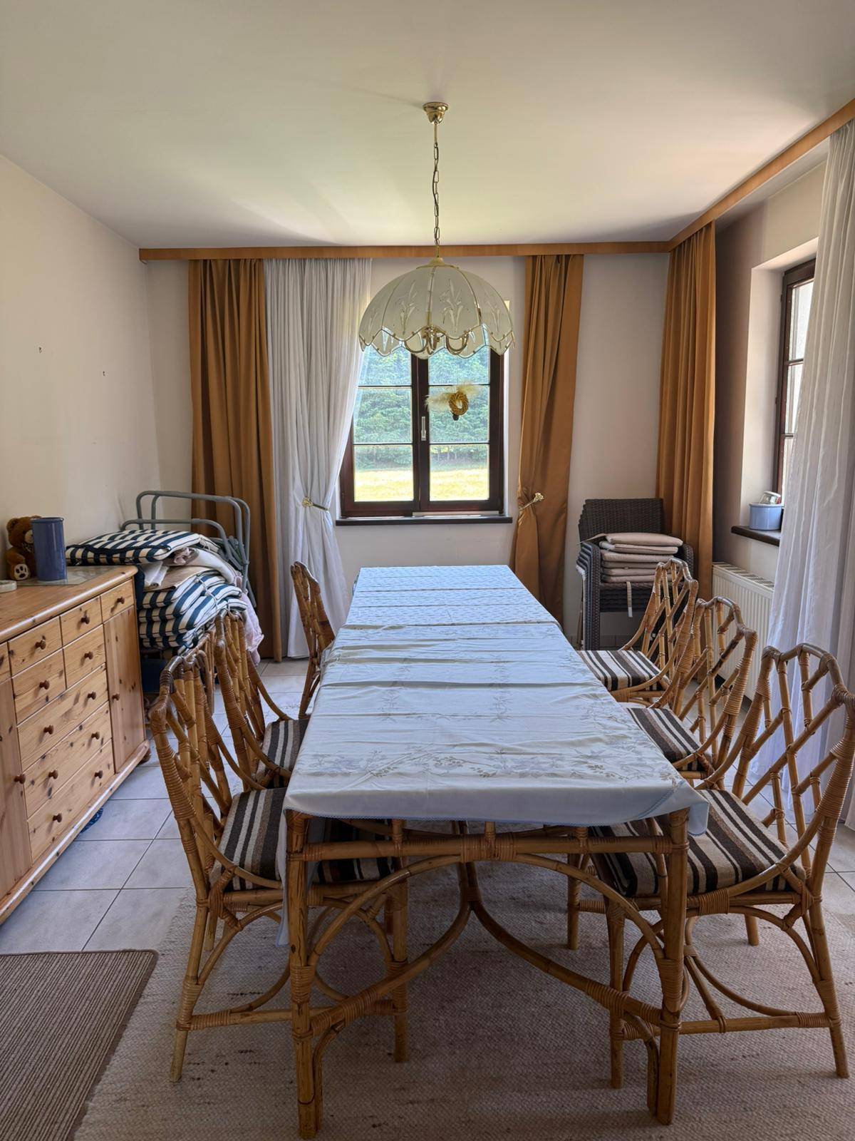 Rustikaler Essbereich mit großem Holztisch, Korbstühlen und einem Bett in der Ecke, auf Fliesenboden.