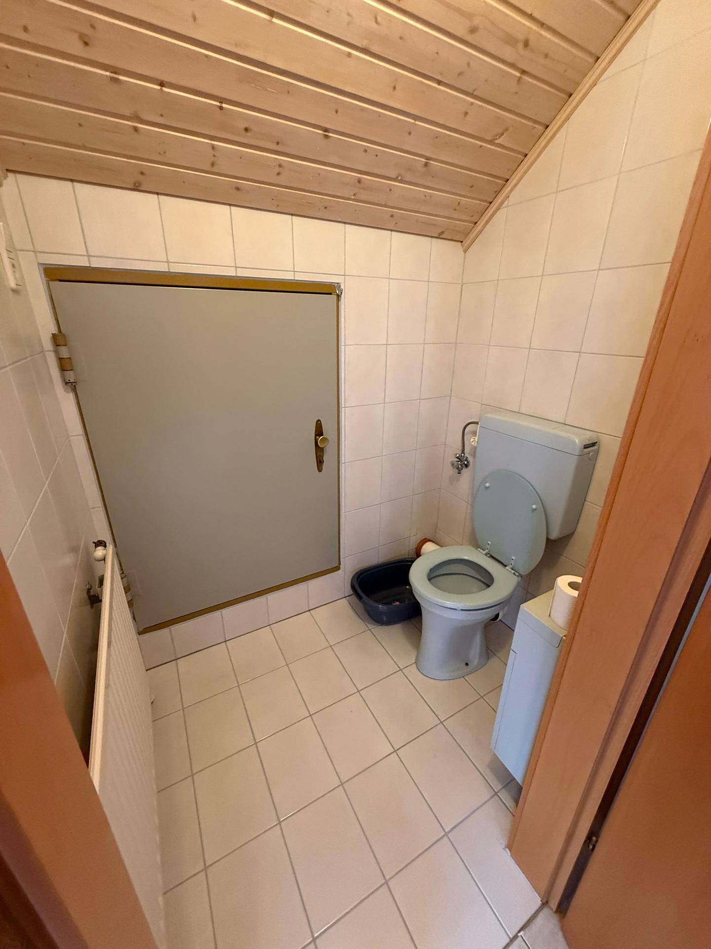 Kleines WC mit hellblauer Toilette, gefliesten Wänden und Holzdachschräge, einfach ausgestattet.