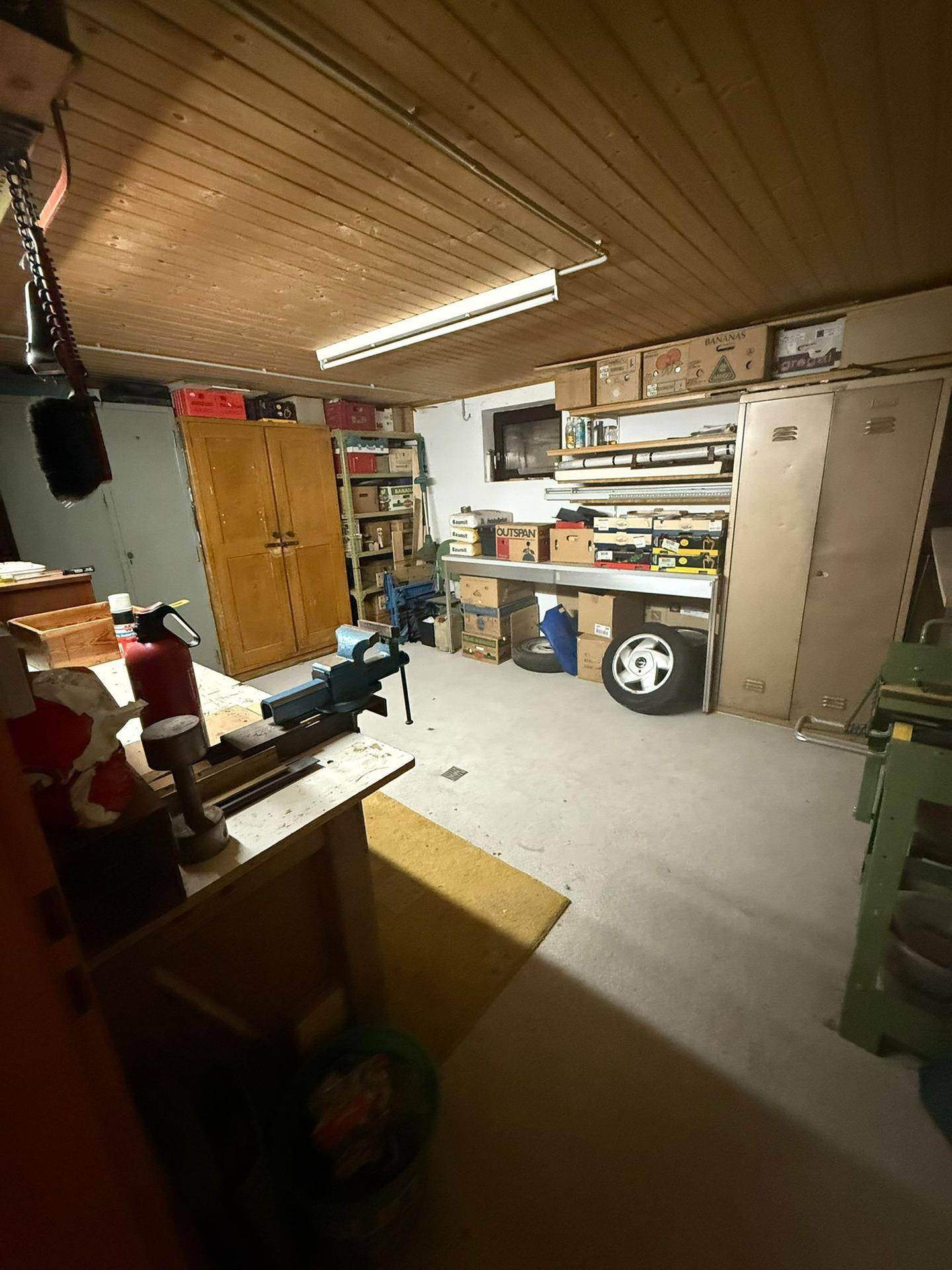 Werkstatt oder Abstellraum im Keller mit Werkbänken, Regalen und Stauraum, unter einer Holzdecke.